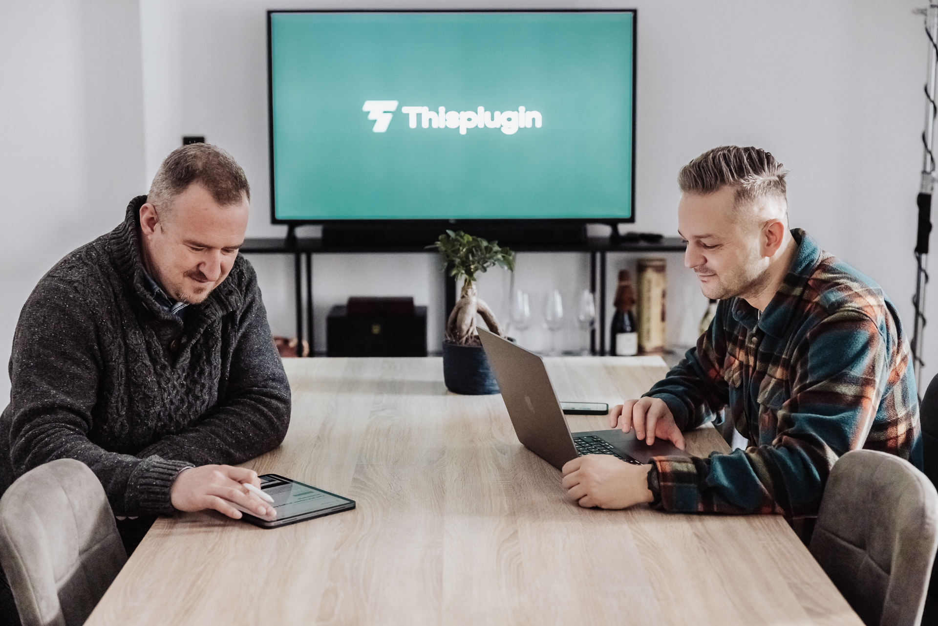 2 mladí muži pracujú na mobilných zariadeniach pri stole v zasadačke, v pozadí TV s logom Thisplugin